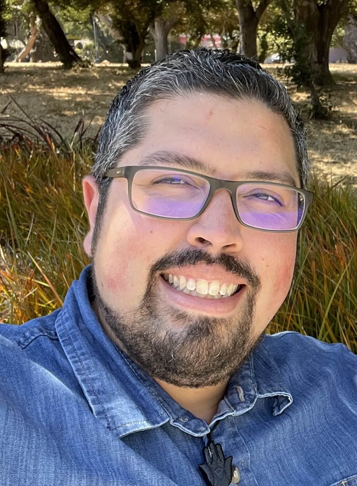 Image features Jaime in the forefront wearing a blue jean shirt, a forrest in the backdrop. 