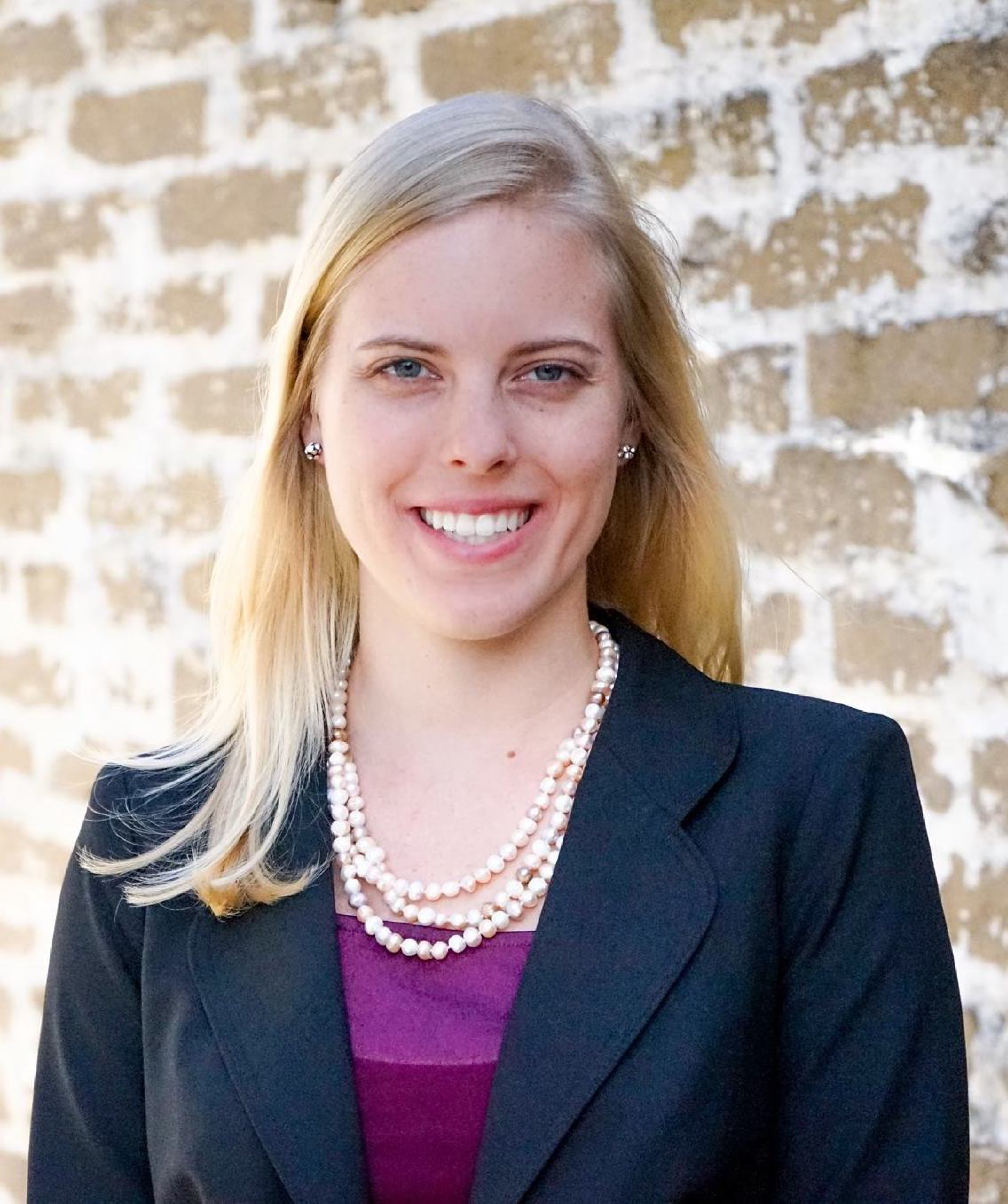 Katy is seen wearing a suit jacket over a purple blouse. A brick backdrop is seen behind her.